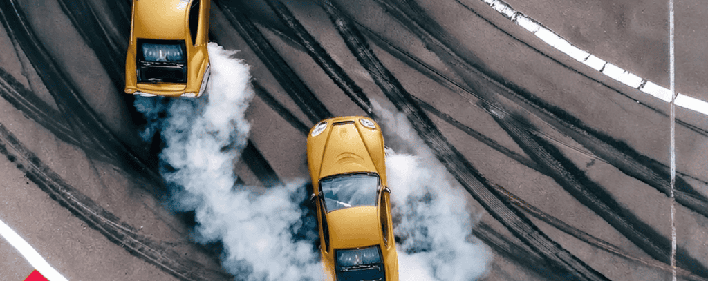 Two yellow cars drifting on a racetrack, creating tire smoke and leaving curved skid marks on the asphalt. The image is taken from above, capturing the motion and patterns made by the drifting cars.
