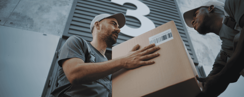 Two delivery workers in gray uniforms and hats handle a cardboard box with a barcode, standing outside near a building with a large number 3 on the wall.