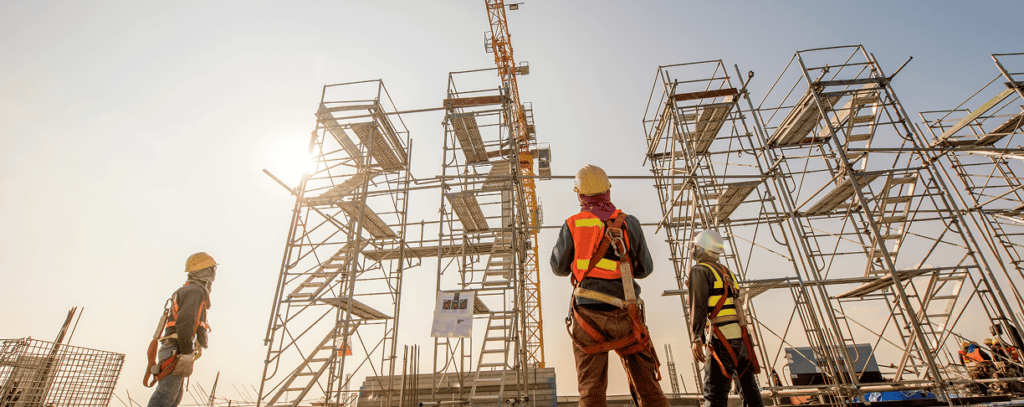 Three construction workers in safety gear stand near tall scaffolding structures on a building site, looking up as the sun sets in the background.