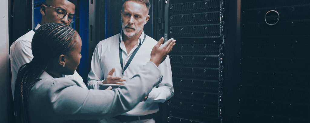 Three people stand in a server room having a discussion. One woman gestures towards the server racks while two men listen attentively, all dressed in professional attire.