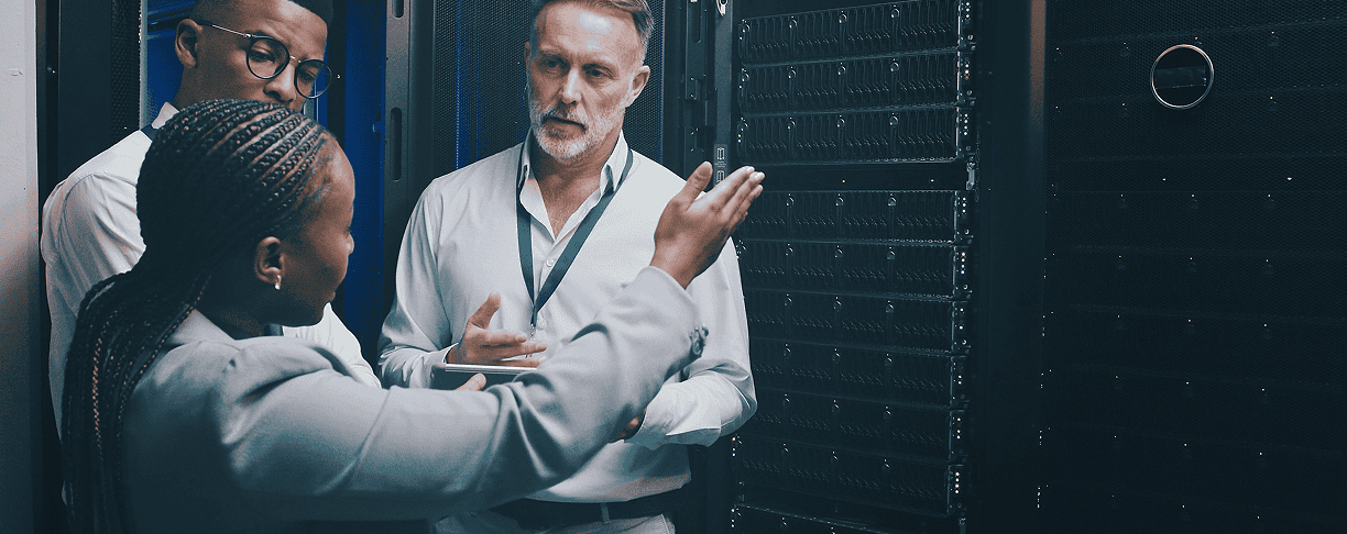Three people stand in a server room having a discussion. One woman gestures towards the server racks while two men listen attentively, all dressed in professional attire.
