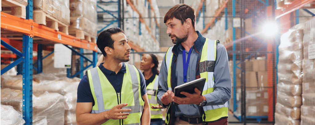 Two workers in reflective vests talk in a warehouse filled with shelves and boxes. One holds a tablet, and a third person is in the background. Bright lighting highlights the organized storage area.