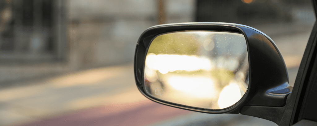 A close-up of a car’s side mirror reflecting sunlight, with a blurred view of a sidewalk, street, and fence in the background.