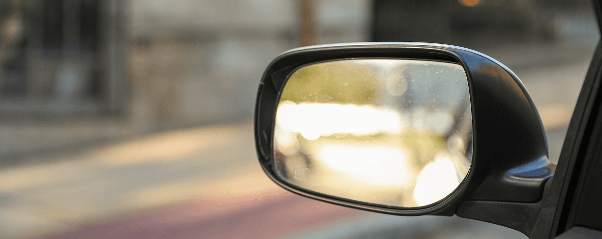 A close-up of a car’s side mirror reflecting sunlight, with a blurred view of a sidewalk, street, and fence in the background.