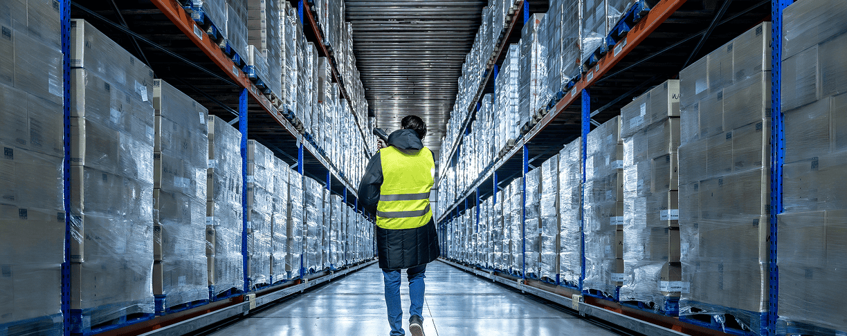 A person wearing a yellow safety vest walks down an aisle in a large warehouse filled with stacked pallets and boxes on high shelves.