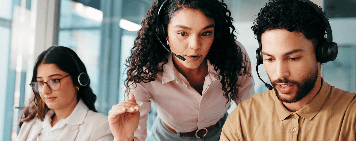 Three people wearing headsets work together in an office. A woman in the center is leaning over to assist a man at a computer, while another woman focuses on her screen.