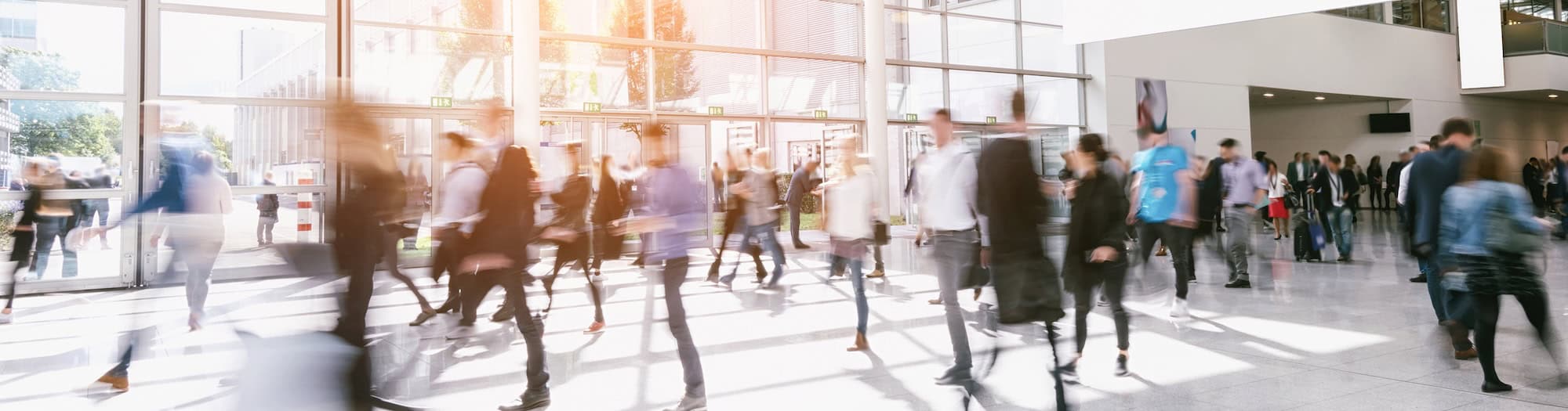 A busy indoor scene shows people walking through a large, bright, modern lobby with glass walls and sunlight streaming in, creating a sense of movement and activity.