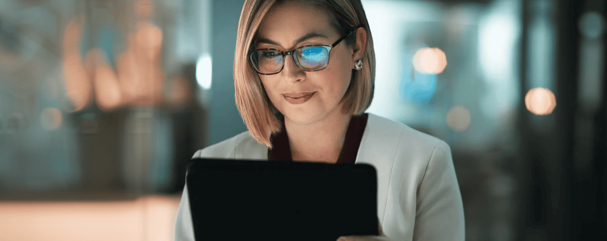 A woman with glasses and a white blazer looks at a laptop screen in a dimly lit, modern office environment.