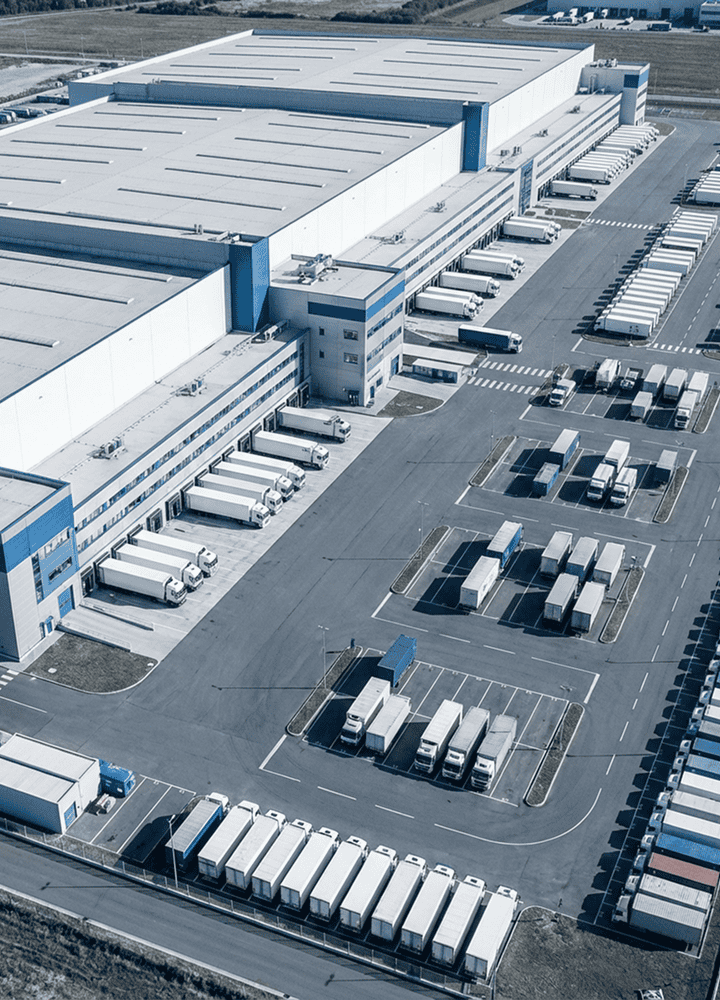Aerial view of a large warehouse with multiple loading docks, several trucks parked and lined up in rows, and trailers in a spacious parking lot. The scene suggests a busy logistics or distribution center.