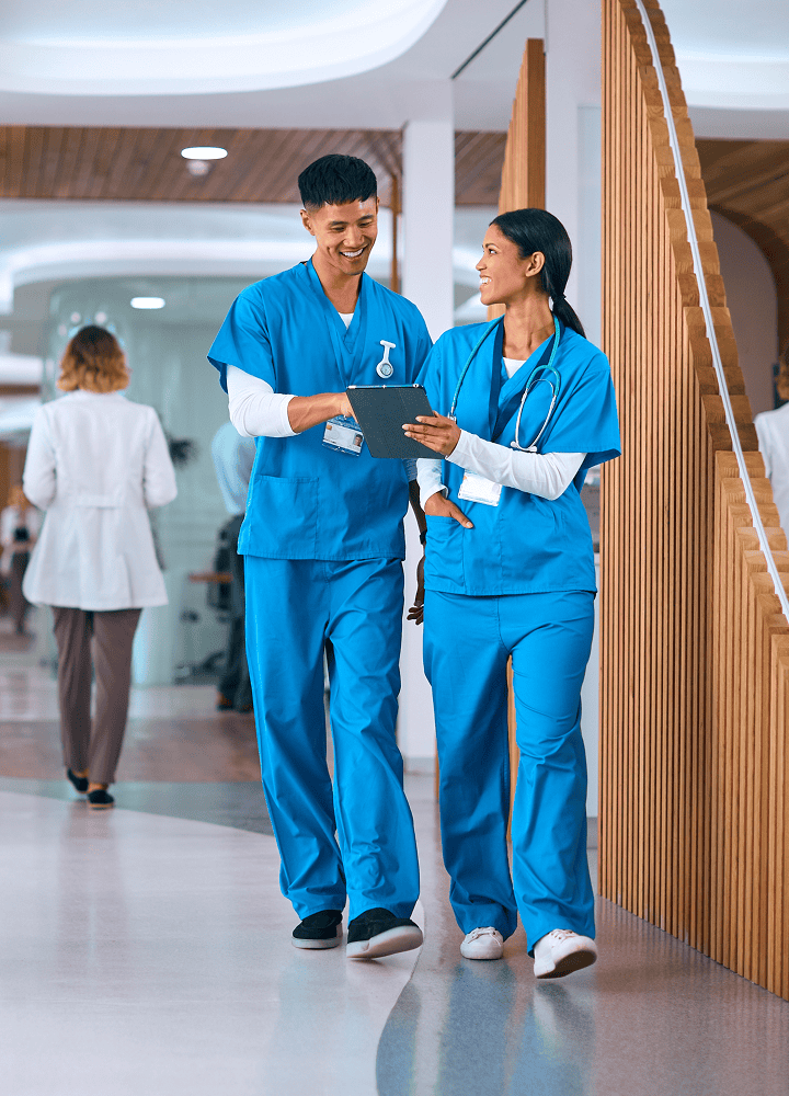 Two healthcare professionals in blue scrubs walk through a hospital hallway, smiling and looking at a tablet together. A person in a white coat is visible in the background.