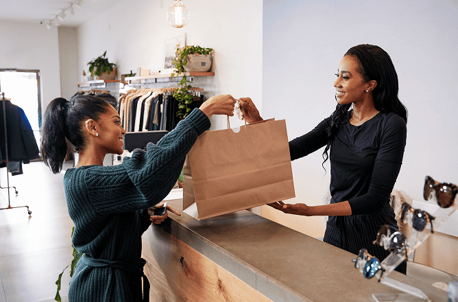 A smiling shop assistant hands a large paper shopping bag to a smiling customer across a counter in a stylish clothing store. Clothing racks, plants, and sunglasses are visible in the background.