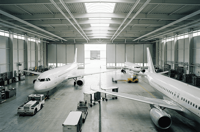 Two commercial airplanes are parked inside a spacious, well-lit aircraft hangar. Maintenance vehicles and equipment are positioned around the planes, and the hangar doors are open to the outside.