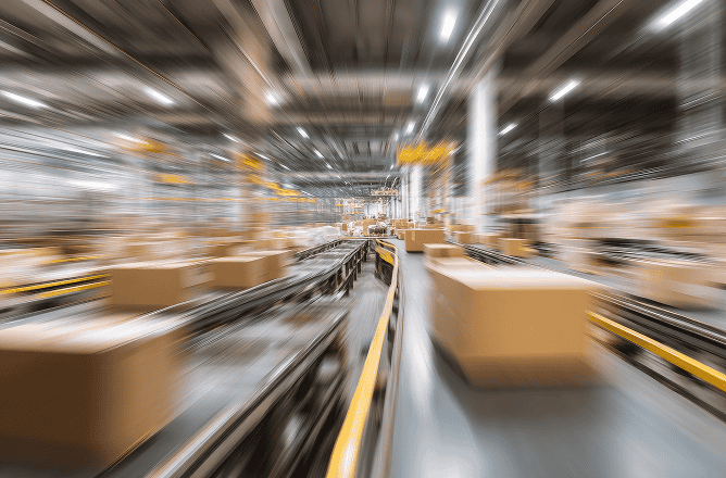 A motion-blurred view of a busy warehouse with cardboard boxes moving quickly along conveyor belts, indicating fast-paced logistics and automated packaging processes.