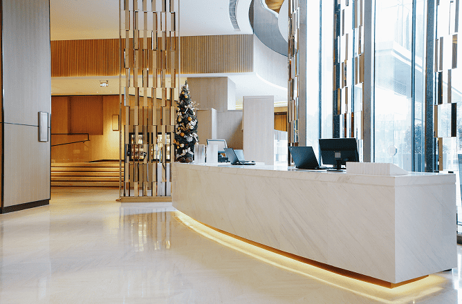 Modern hotel lobby with a white marble reception desk, two computer monitors, decorative metal dividers, a lit Christmas tree in the background, and large floor-to-ceiling windows letting in natural light.