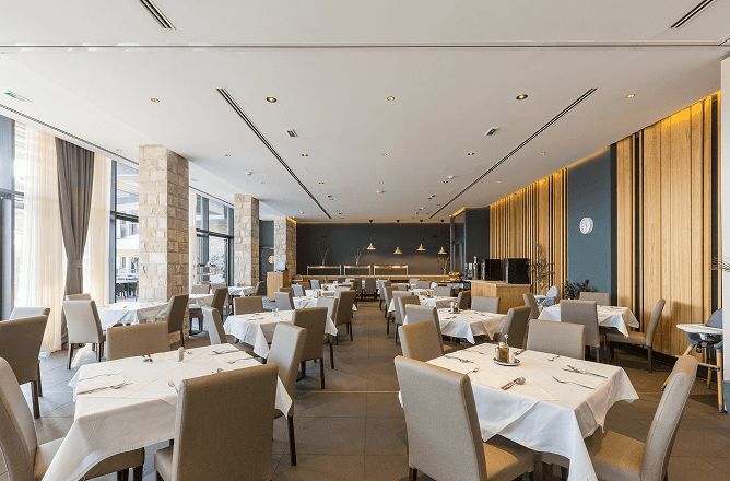 Modern restaurant dining area with neatly arranged tables covered in white cloths, beige chairs, large windows letting in natural light, and minimalist decor with wood paneling and stone columns.