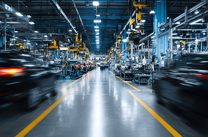 A wide view of an automotive factory with assembly lines, machinery, and robotic arms. Blurred cars move along the lines, and bright lights illuminate the spacious, modern industrial setting.
