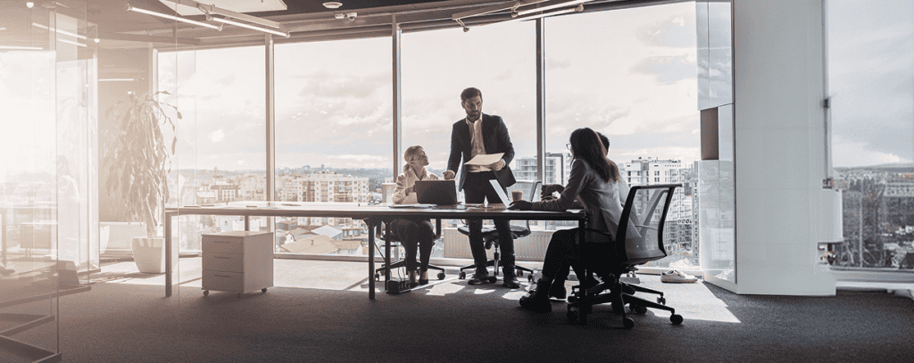 Three people in business attire meet around a table in a modern office with large windows, cityscape views, and sunlight streaming in.