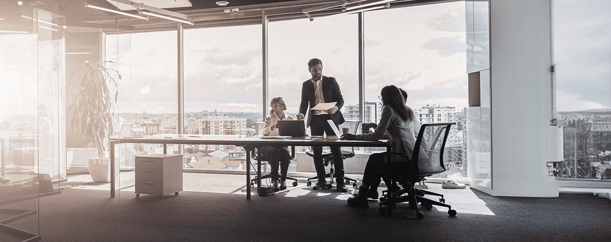 Three people in business attire meet around a table in a modern office with large windows, cityscape views, and sunlight streaming in.