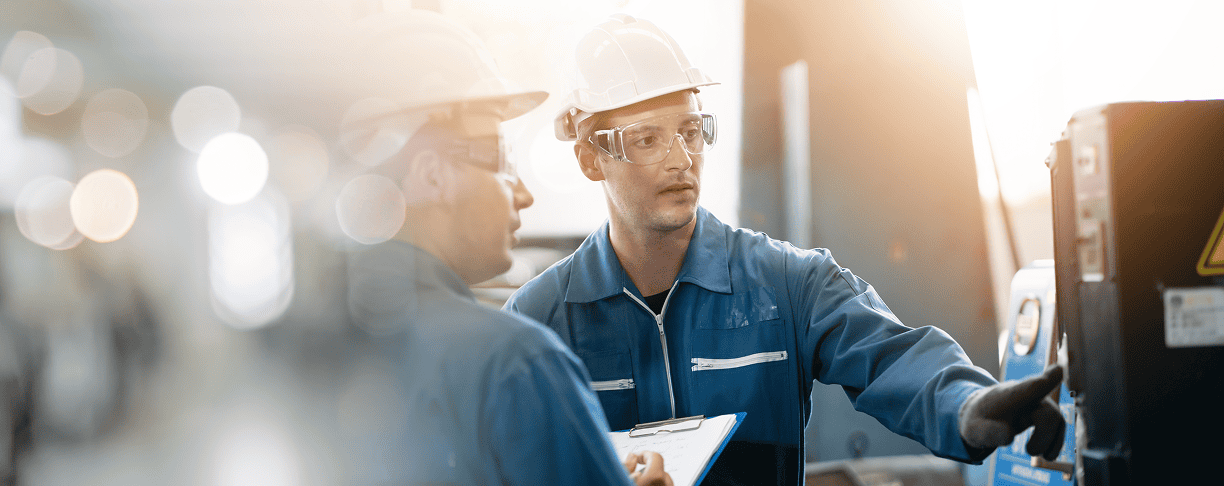 Two engineers wearing hard hats, safety glasses, and blue coveralls discuss machinery, with one holding a clipboard and the other pointing at equipment in an industrial setting.