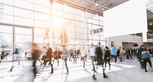 A busy, sunlit modern lobby with large windows, high ceilings, and many people walking or standing, creating a blurred effect from their movement. A large blank white sign hangs on the wall above.
