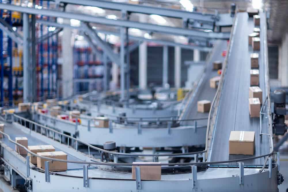 Boxes move along a network of automated conveyor belts in a large warehouse or distribution center, with shelves of goods visible in the background.
