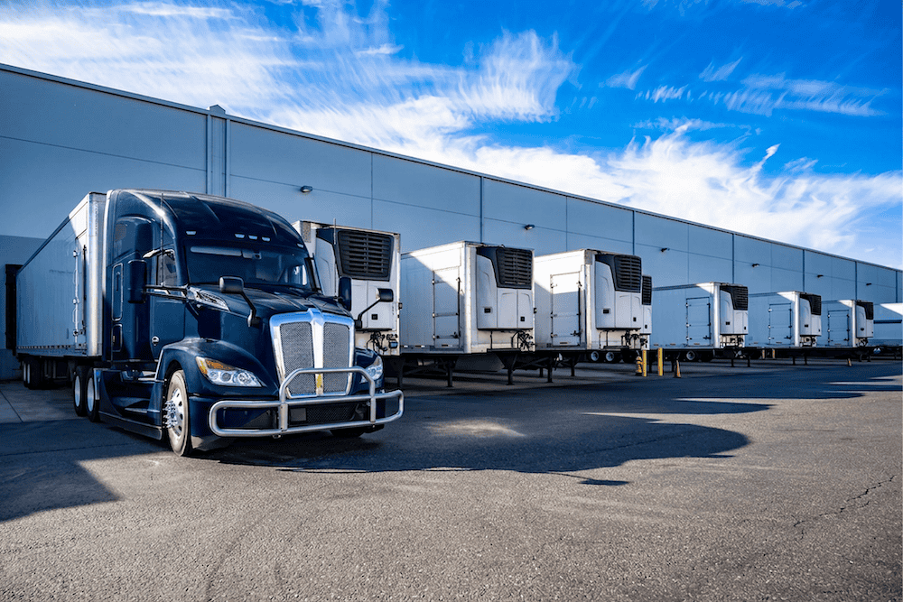A row of white semi-trailers is lined up at loading docks outside a large warehouse, with a blue semi-truck parked in the foreground under a blue sky with wispy clouds.