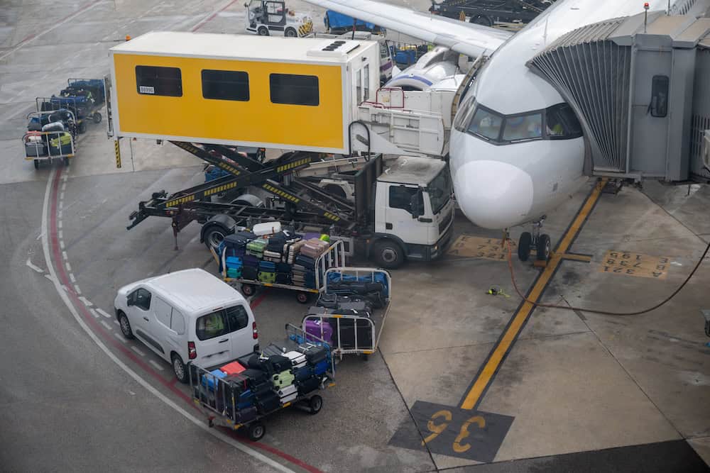 A passenger airplane is parked at a gate with a yellow boarding bridge, while luggage carts and ground vehicles load and unload suitcases onto the plane on a rainy airport tarmac.